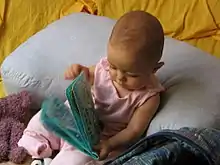 A baby sits a rug on the floor surrounded by toys. He stares down at a cloth book in his hands in a posture of interest and concentration.