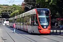 A red and silver electrified tram running through a street as a crossing pedestrian waits for it to pass