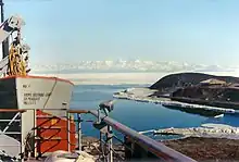  In left foreground are the upper works of a modern ship. To the right, on a low headland fringed with sea ice, isa square hut with a pitched roof. In te distance is a range of icy mountains