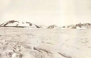  A mountain with a rounded summit and snowy slopes, with smaller hills to its right, rising from flat snow-covered ground