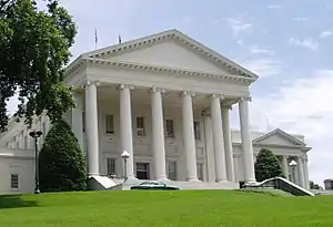 All white Neoclassical building with pediment and six columns rises on a grassy hill with a large American elm tree in the left foreground. Two boxier, but similarly styled wings are attached at the building's rear.