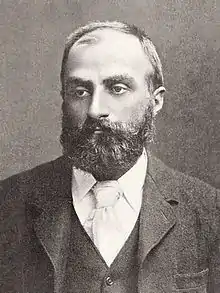  Heavily bearded man with dark receding hair, wearing a dark coloured jacket, white collar and pale tie. He is looking slightly to the left, with a solemn expression