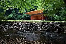 A small, boxy, wooden stage with a trapezoidal overhang stands in the center of meadow. In the foreground is a running stream with a stone embankment.